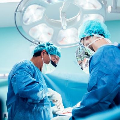 three people in blue gowns, surgical masks and gloves stand under a bright light looking down